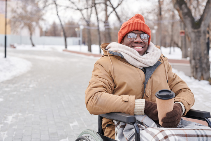 Man in warm clothing sitting in a manual wheelchair holding a cup of coffee with snow in the background.