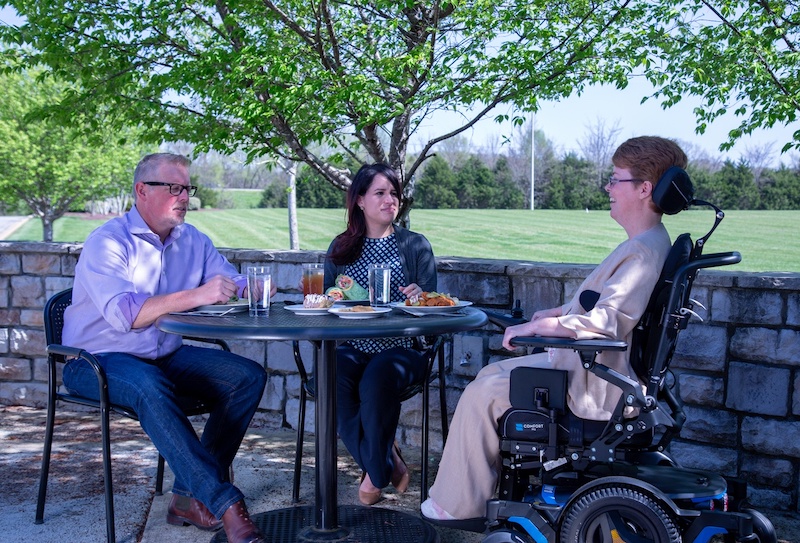 Three people sit around a table enjoying the outdoors, one in a Permobil powerchair with headrest