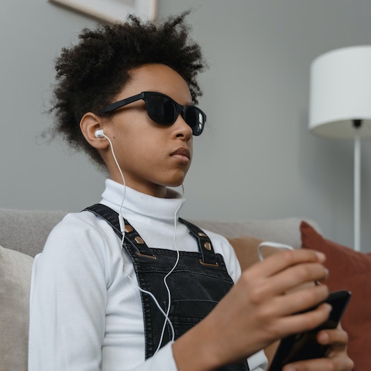 Blind child wearing sunglasses and ear buds plugged into a smartphone.